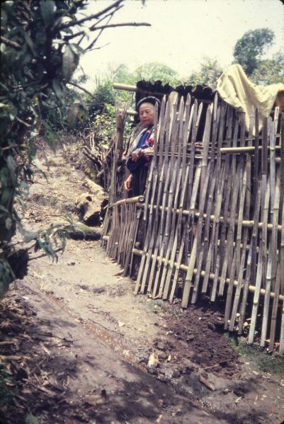 An old Lisu woman is standing behind a bamboo fence. EFEO_KLEO00776